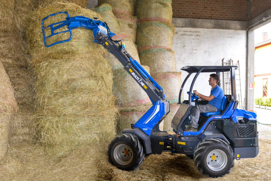 man driving a mini loader and using an hay bale grapple to grab a hay bale, seen from the left side